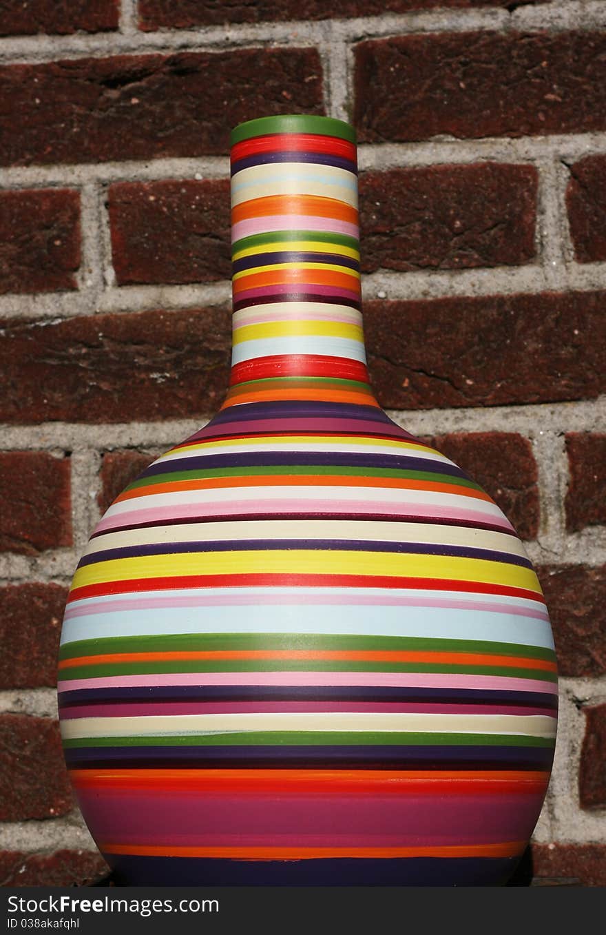 A mulit-colored vase is set against a brick wall. The vase is full of color, including green, red, purple, yellow, white, blue, orange and pink. A mulit-colored vase is set against a brick wall. The vase is full of color, including green, red, purple, yellow, white, blue, orange and pink.