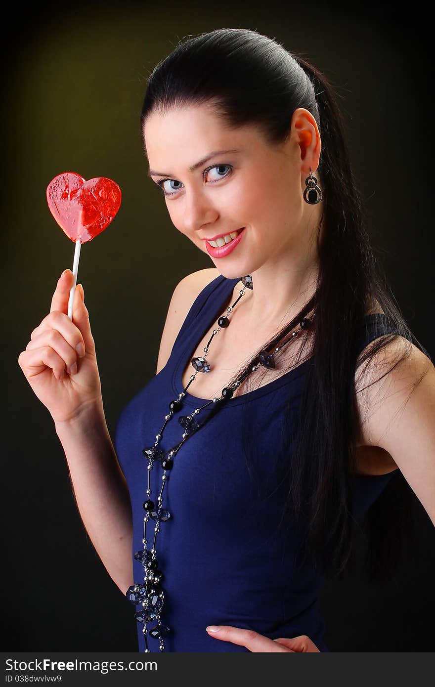 Portrait beautiful young sexy brunette girl with red lollipop heart. On a black background.