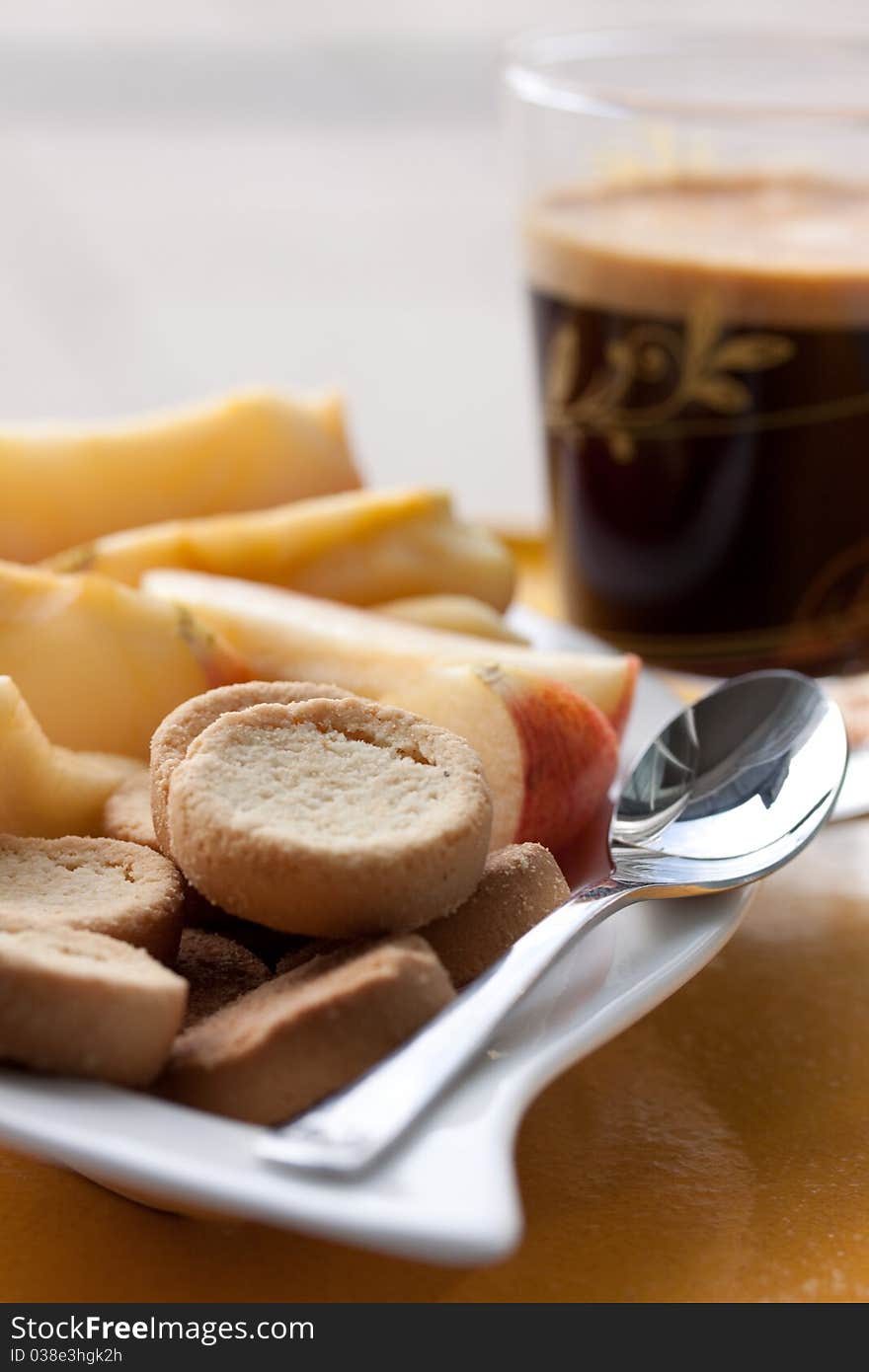 This is a small white plate with cookies and apple slices for a small snack and a glas cup of coffee. This is a small white plate with cookies and apple slices for a small snack and a glas cup of coffee