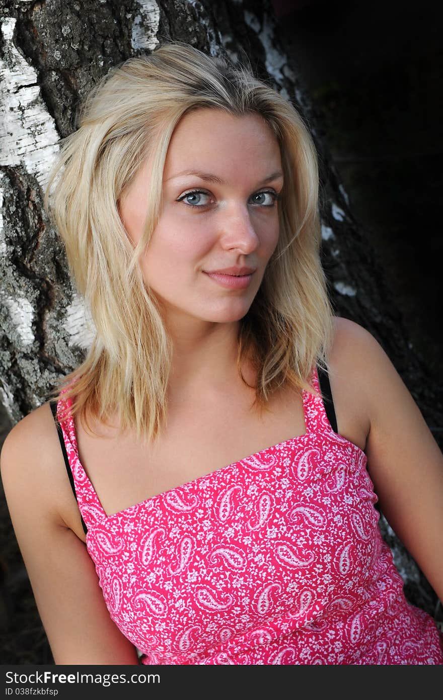 Portrait of a beautiful blonde woman with long hair and a smile near tree in pink dress. Portrait of a beautiful blonde woman with long hair and a smile near tree in pink dress