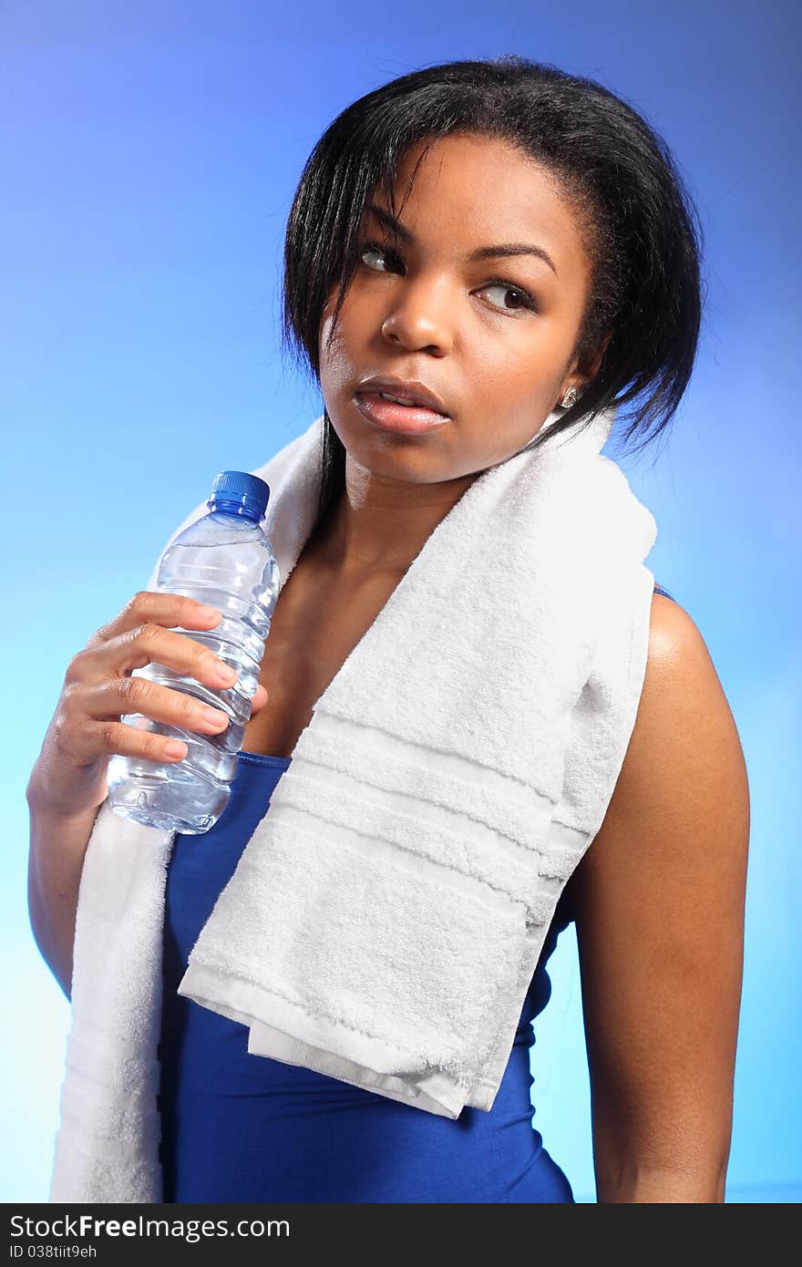 Portrait of beautiful black woman, looking tired after working out. She is wearing a blue vest, has a white towel around her shoulders and holding a bottle of water. Portrait of beautiful black woman, looking tired after working out. She is wearing a blue vest, has a white towel around her shoulders and holding a bottle of water.