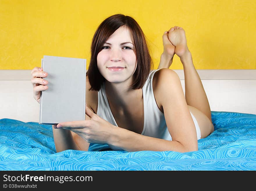 Young woman in white undergarment lying in bed on belly and showing book, bright interior. Young woman in white undergarment lying in bed on belly and showing book, bright interior