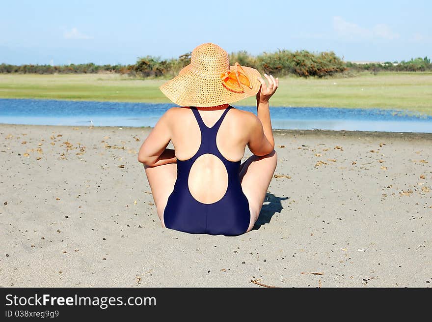 Woman in dark blue bikini sits on hot sand in a yellow hat to river bank