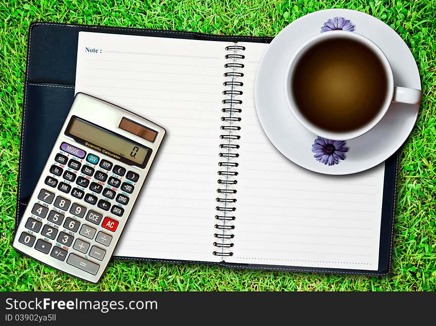 White cup of coffee and blank notebook with calculator on fresh green grass background. White cup of coffee and blank notebook with calculator on fresh green grass background