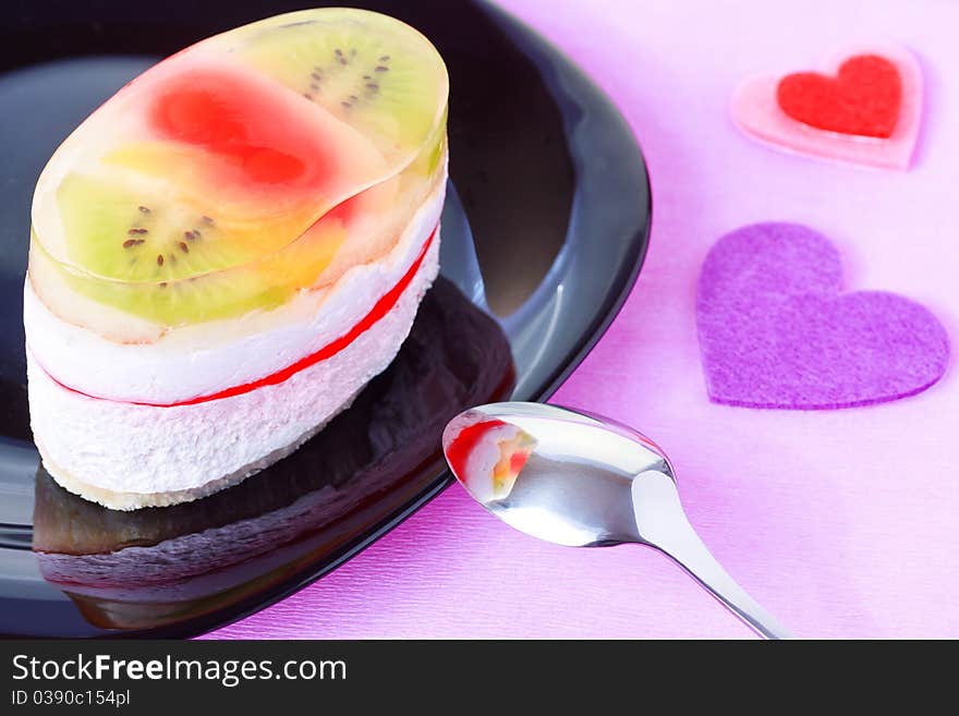 Closeup of cake with jelly and fruits on a black plate with Valentine`s day heart shaped decorations on a purple background. Closeup of cake with jelly and fruits on a black plate with Valentine`s day heart shaped decorations on a purple background