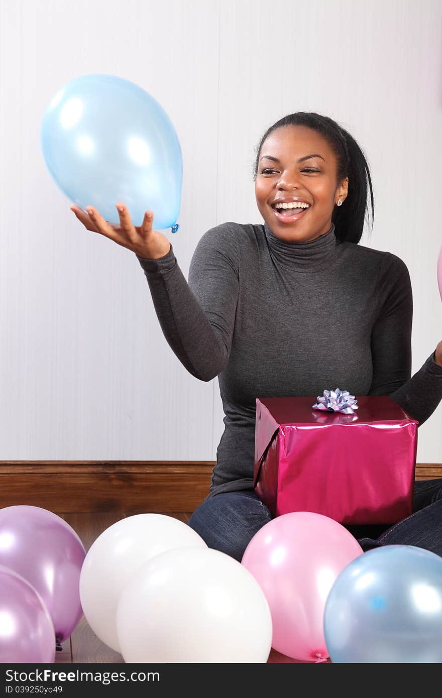 Balloons and presents. Time to celebrate for beautiful, smiling young african american girl, sitting on the wood floor at home, with a birthday present, surrounded by balloons. Balloons and presents. Time to celebrate for beautiful, smiling young african american girl, sitting on the wood floor at home, with a birthday present, surrounded by balloons.