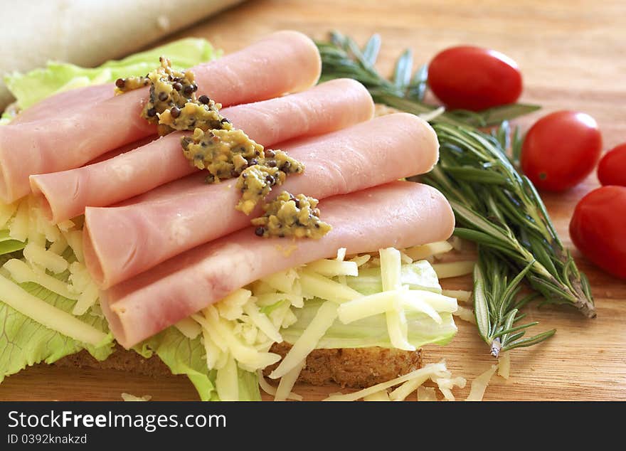 Tasty open sandwich with green lettuce, grated cheese, smoked ham and wholegrain mustard on wholewheat bread with rosemary and tomatoes on chopping board. Tasty open sandwich with green lettuce, grated cheese, smoked ham and wholegrain mustard on wholewheat bread with rosemary and tomatoes on chopping board