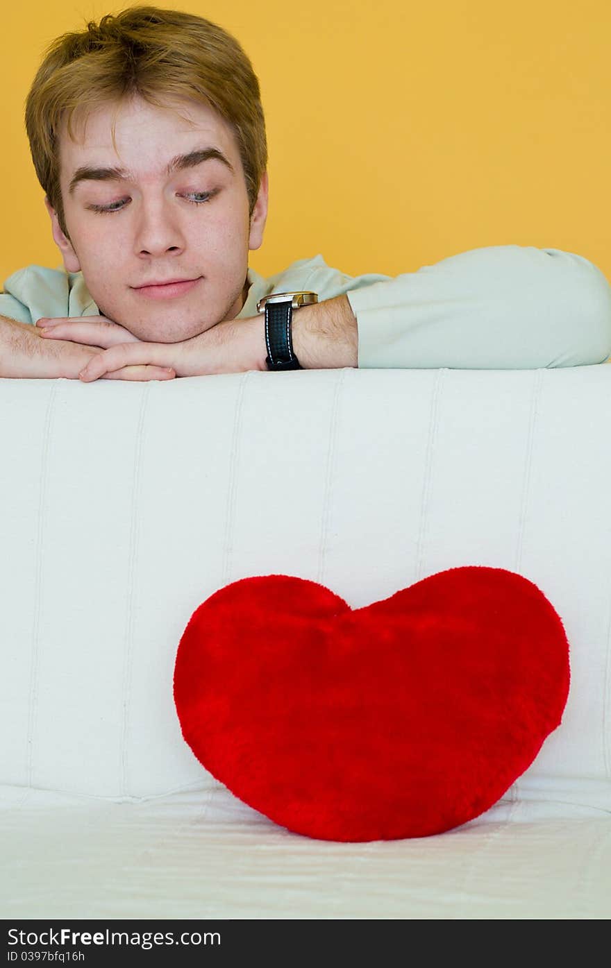 Blond young man looking at a red heart shaped pillow. Blond young man looking at a red heart shaped pillow