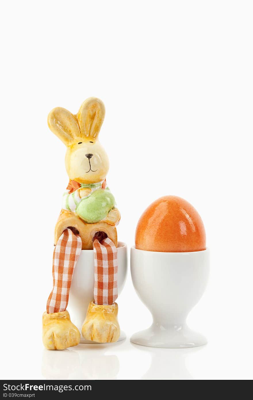 Easter bunny sitting on an egg-cup close to an orange egg in an egg-cup on a white background