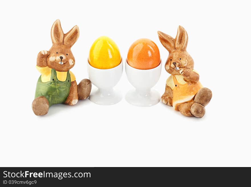 Two easter bunnies sitting close to orange and yellow eggs in egg-cups on a white background. Two easter bunnies sitting close to orange and yellow eggs in egg-cups on a white background