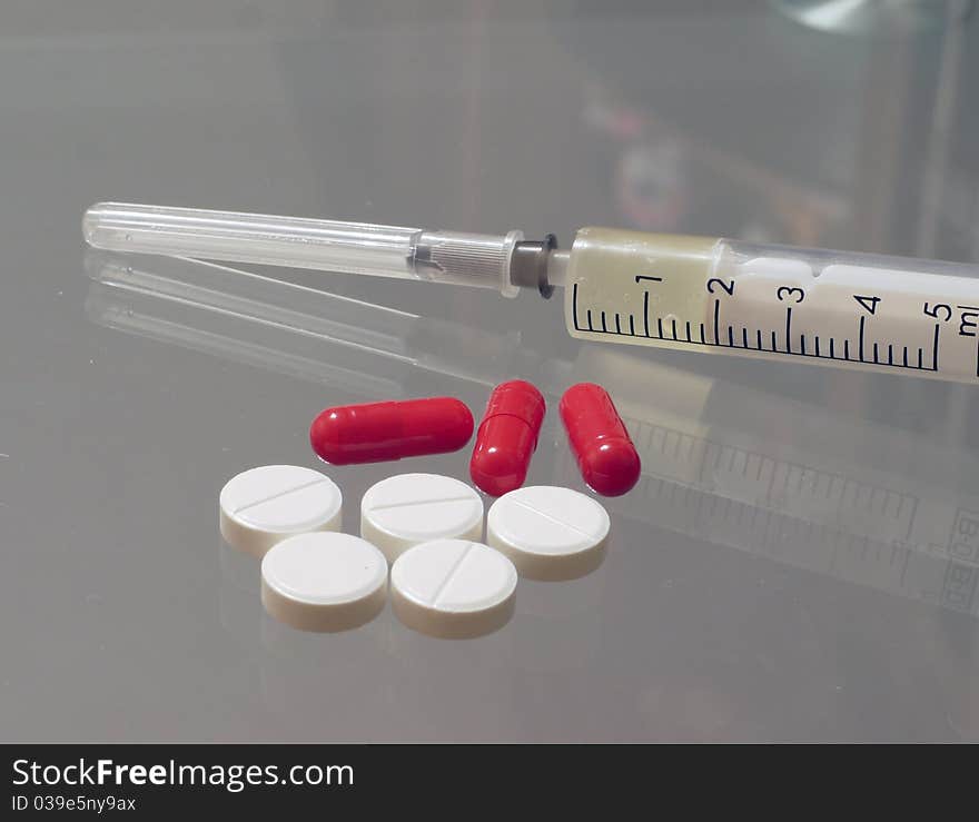 Five white round tablets, three red pills and one syringe lying on a glass table close up. Five white round tablets, three red pills and one syringe lying on a glass table close up