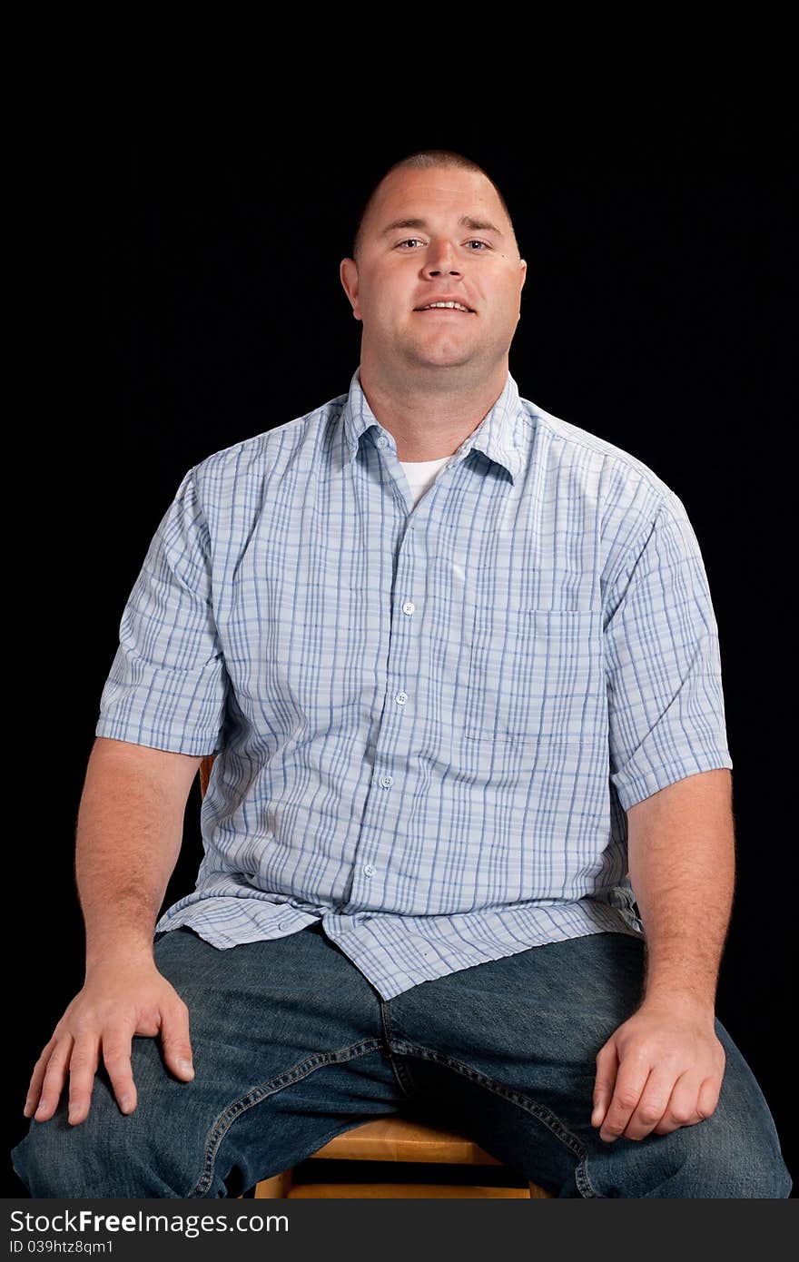 A young man sitting down on a wooden chair.  He has an odd look on his face.  He is wearing a blue shirt and there is a black background. A young man sitting down on a wooden chair.  He has an odd look on his face.  He is wearing a blue shirt and there is a black background.