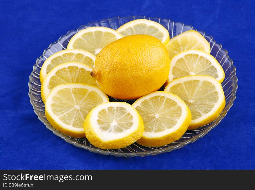 Whole lemon surrounded by lemon slices on a clear glass palte on a bright blue background. Whole lemon surrounded by lemon slices on a clear glass palte on a bright blue background