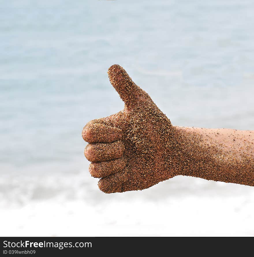 Beach fun with a hand with sand giving a thumbs up, with the ocean beach waves in the background.