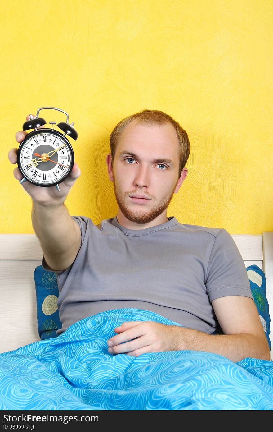 Young man lying in bed and showing alarm clock and looking at camera, bright interior