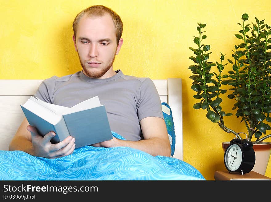 Young man lying in bed and reading book, table with plant and clock, bright interior