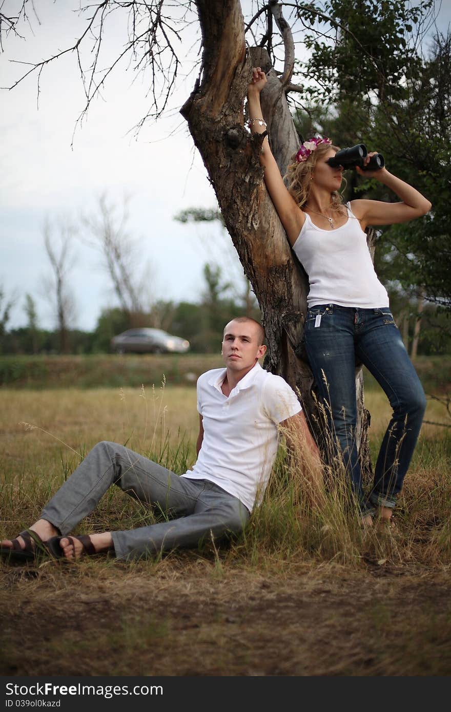 The guy sits near a tree, nearby there is a girl and looks in the field-glass. The guy sits near a tree, nearby there is a girl and looks in the field-glass