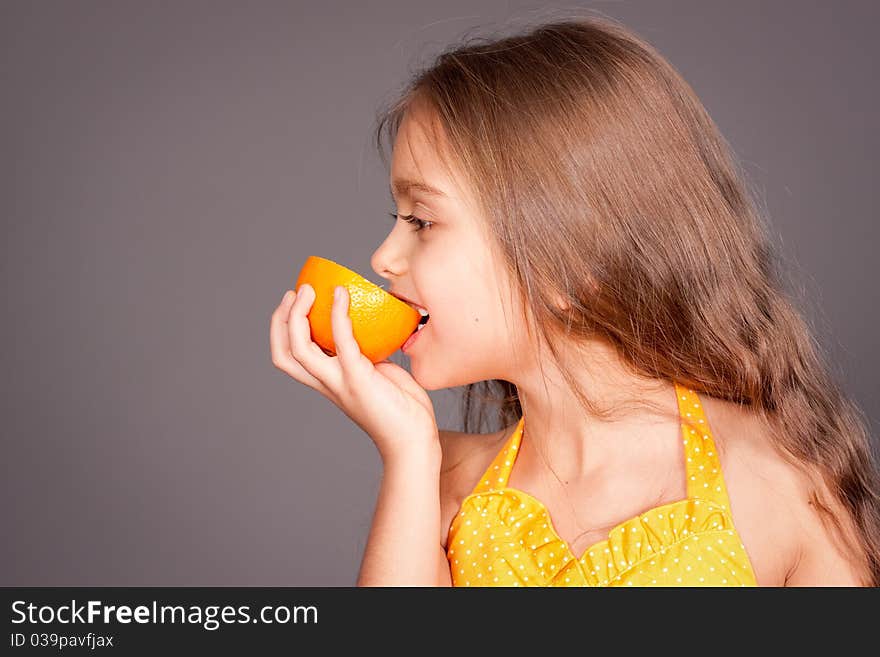 Beautiful young cheerful girl has turned in profile and bites orange, against gray background