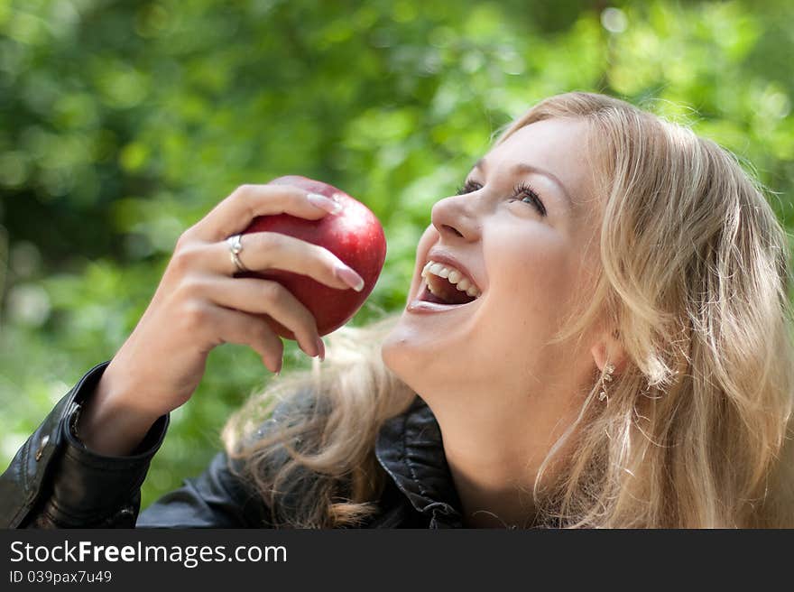 Beautiful young cheerful woman has turned in profile and bites red big apple, against green garden.