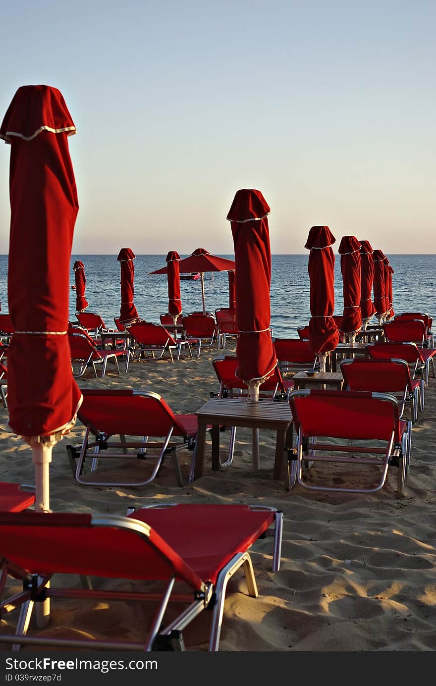 Red sunshades and sunbeds at sandy beach at summer sundown with blue sea and sky in background.