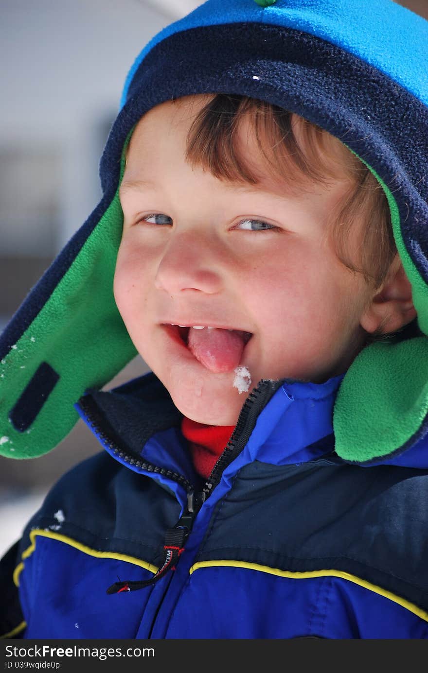 A toddler boy dressed in blue and green hat and coat outside playing in the snow and licking a snowflake off his chin. A toddler boy dressed in blue and green hat and coat outside playing in the snow and licking a snowflake off his chin.