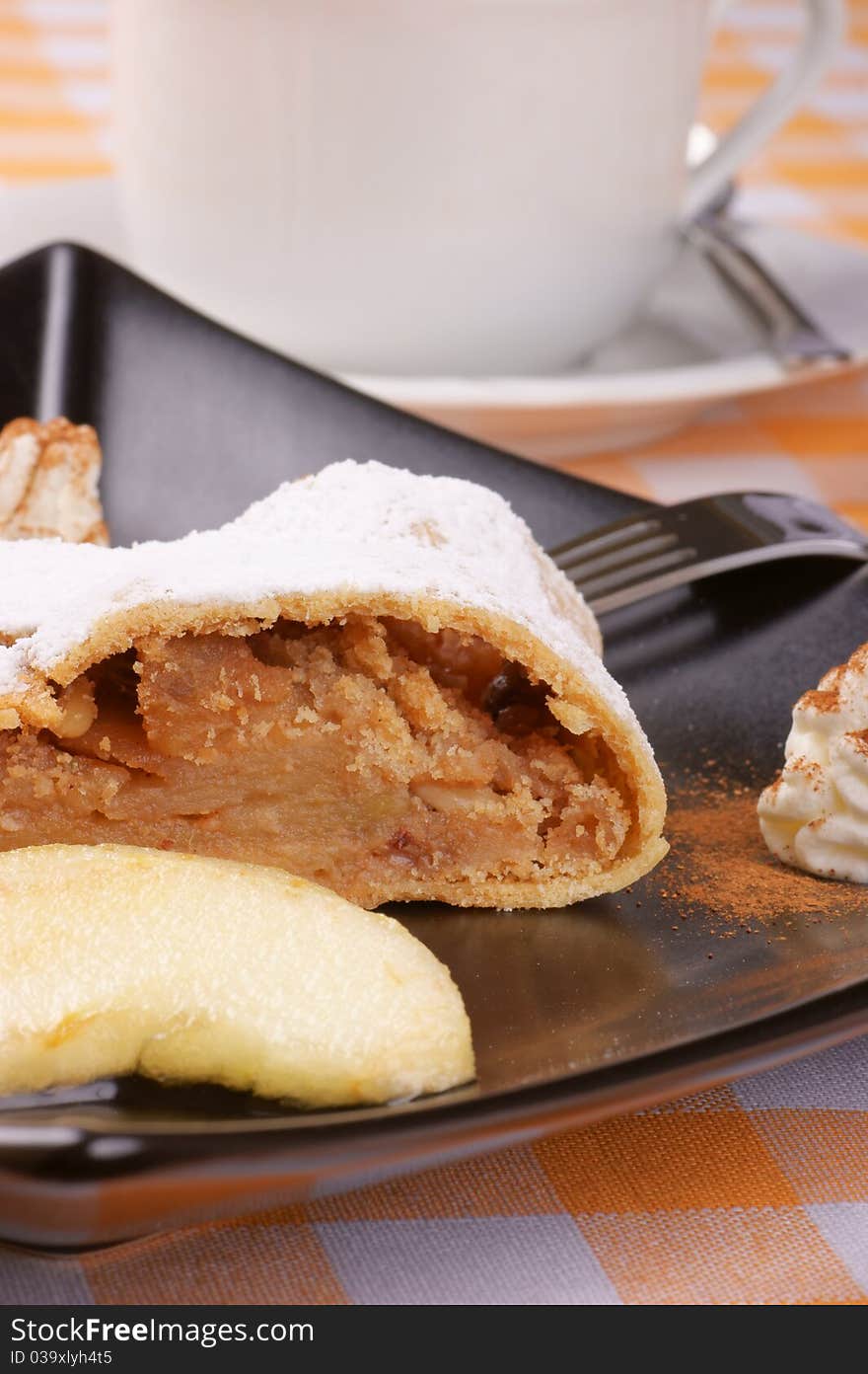 Apple strudel (Apfelstrudel) served on a black plate with a tea cup in the background. Selective focus, shallow DOF.