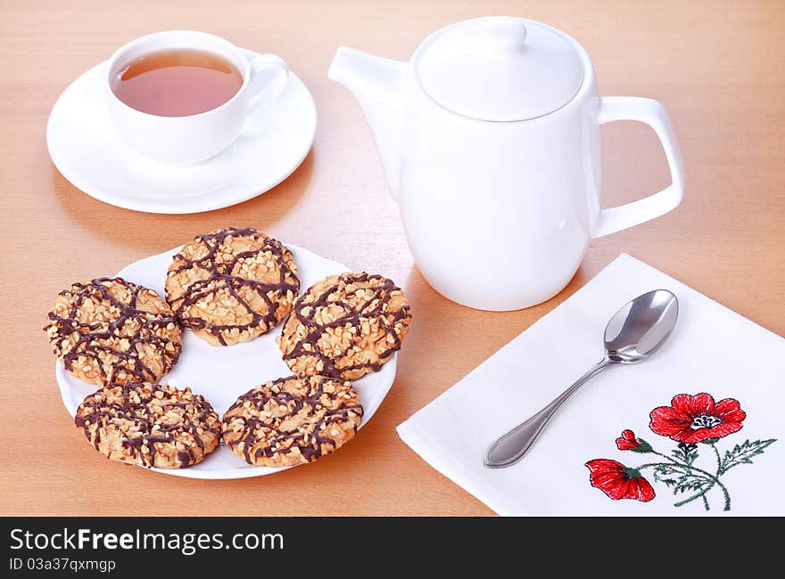 Biscuits with chocolate and peanut decoration on a white plate, teapot and a cup of tea on a brown table background