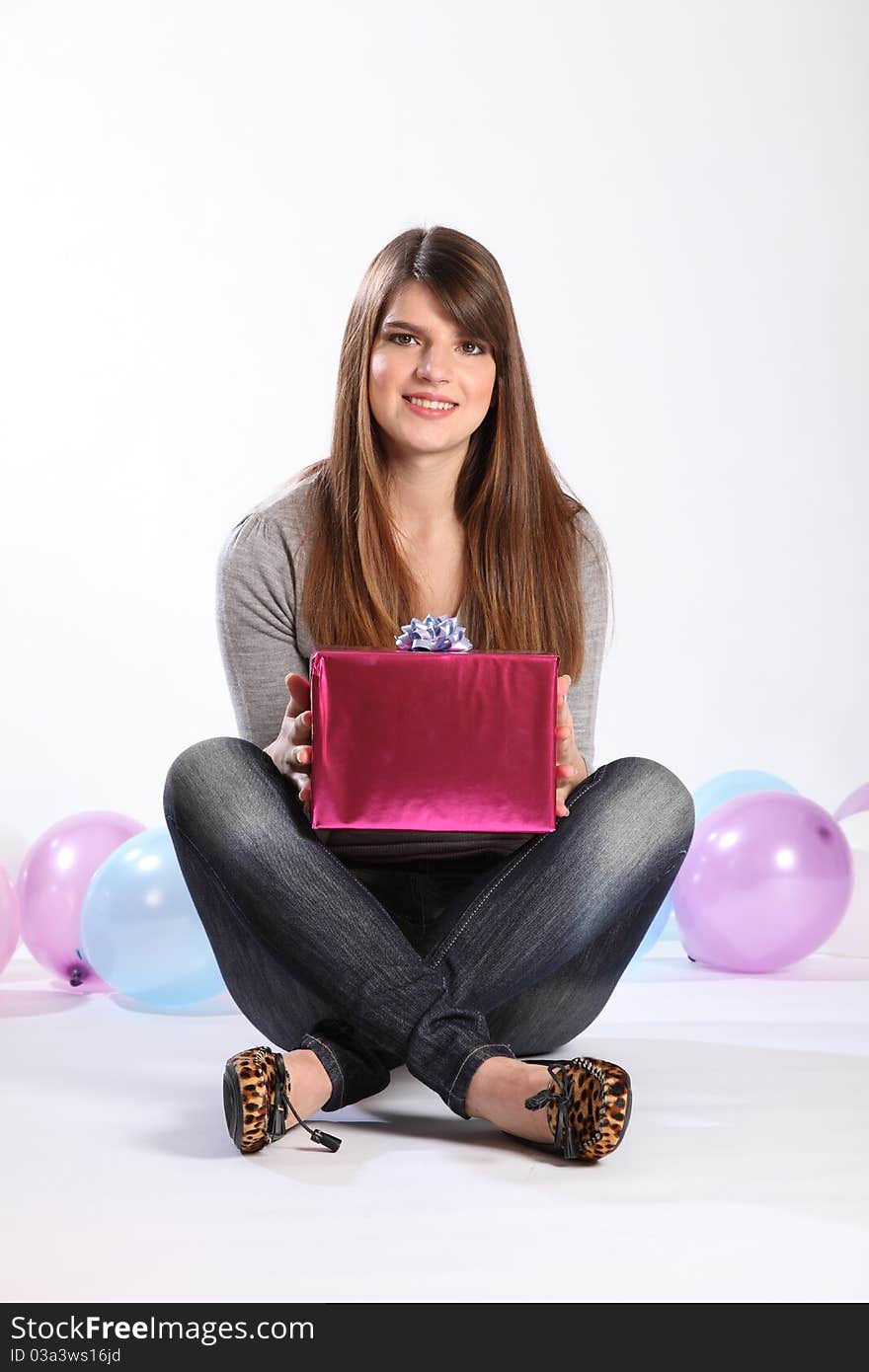 Beautiful young caucasian, student girl with long hair, sitting on the floor with a birthday present. She is wearing a grey top and blue jeans, there are balloons in the background. Beautiful young caucasian, student girl with long hair, sitting on the floor with a birthday present. She is wearing a grey top and blue jeans, there are balloons in the background.