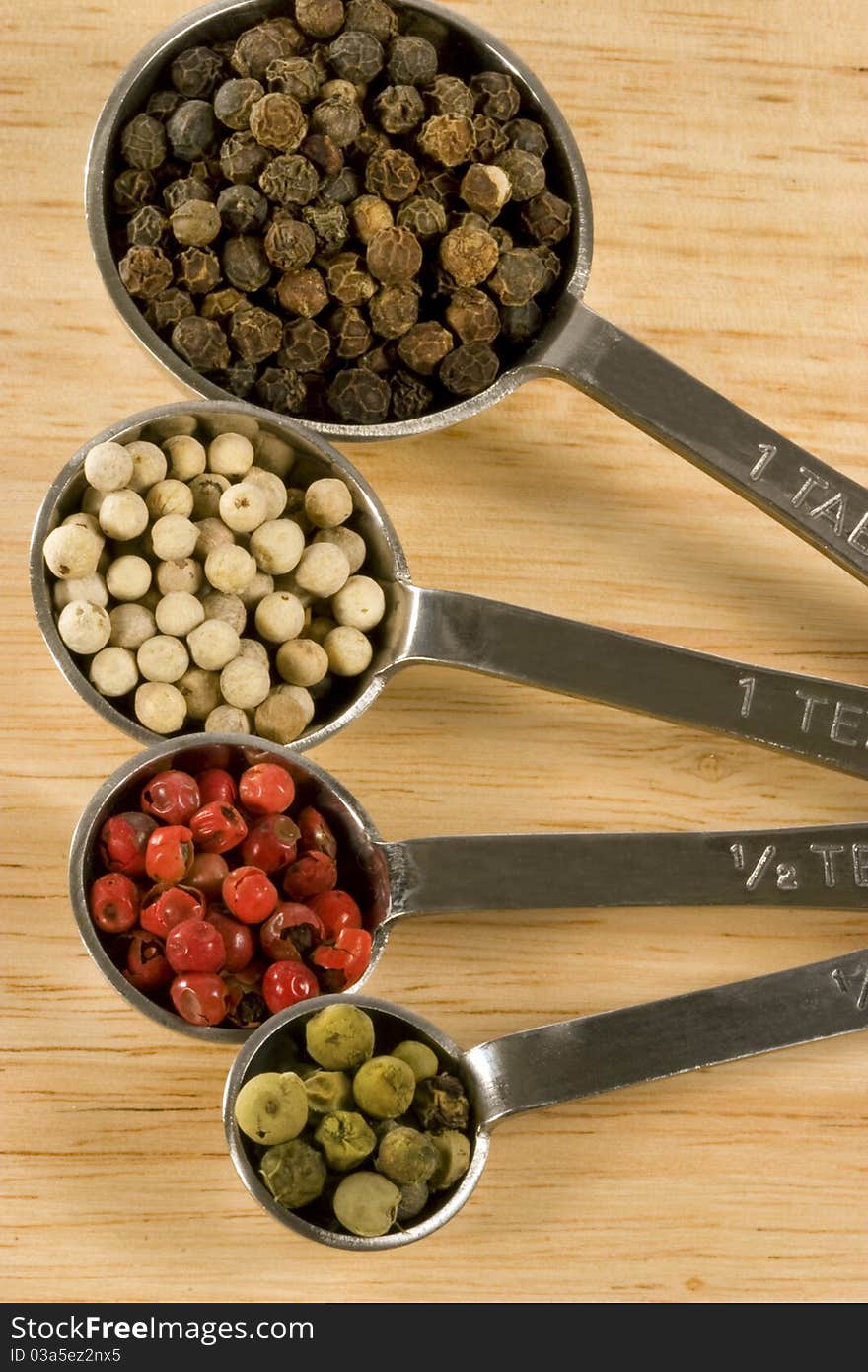 Metal measuring spoons on wood background, each spoon containing different colored whole peppercorns including black, white, red and green. Metal measuring spoons on wood background, each spoon containing different colored whole peppercorns including black, white, red and green