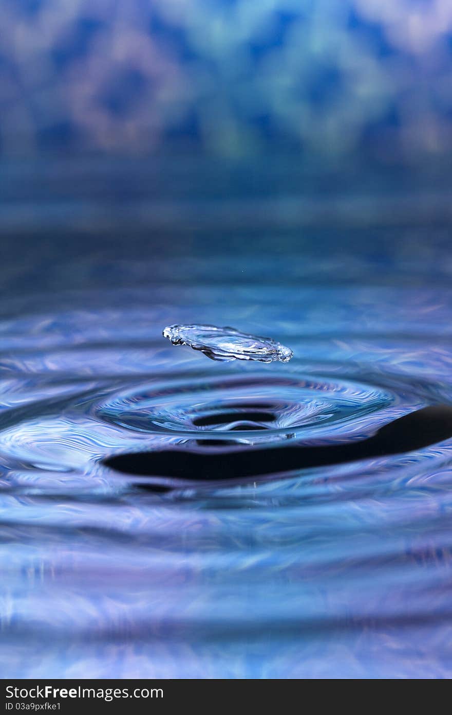 A blue abstract background of a close up of transparent water drops falling in water, making beautifull ripples and flowing waves in the surface and showing the pure energy it creates. Small depth of field.