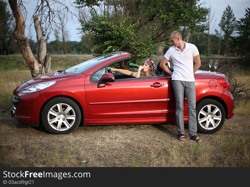 The girl in a red cabriolet drinks champagne, nearby there is a guy. The girl in a red cabriolet drinks champagne, nearby there is a guy