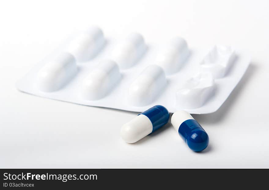 Two blue and white pharmaceutical capsules in front of blister packaging all close up on white. Two blue and white pharmaceutical capsules in front of blister packaging all close up on white.