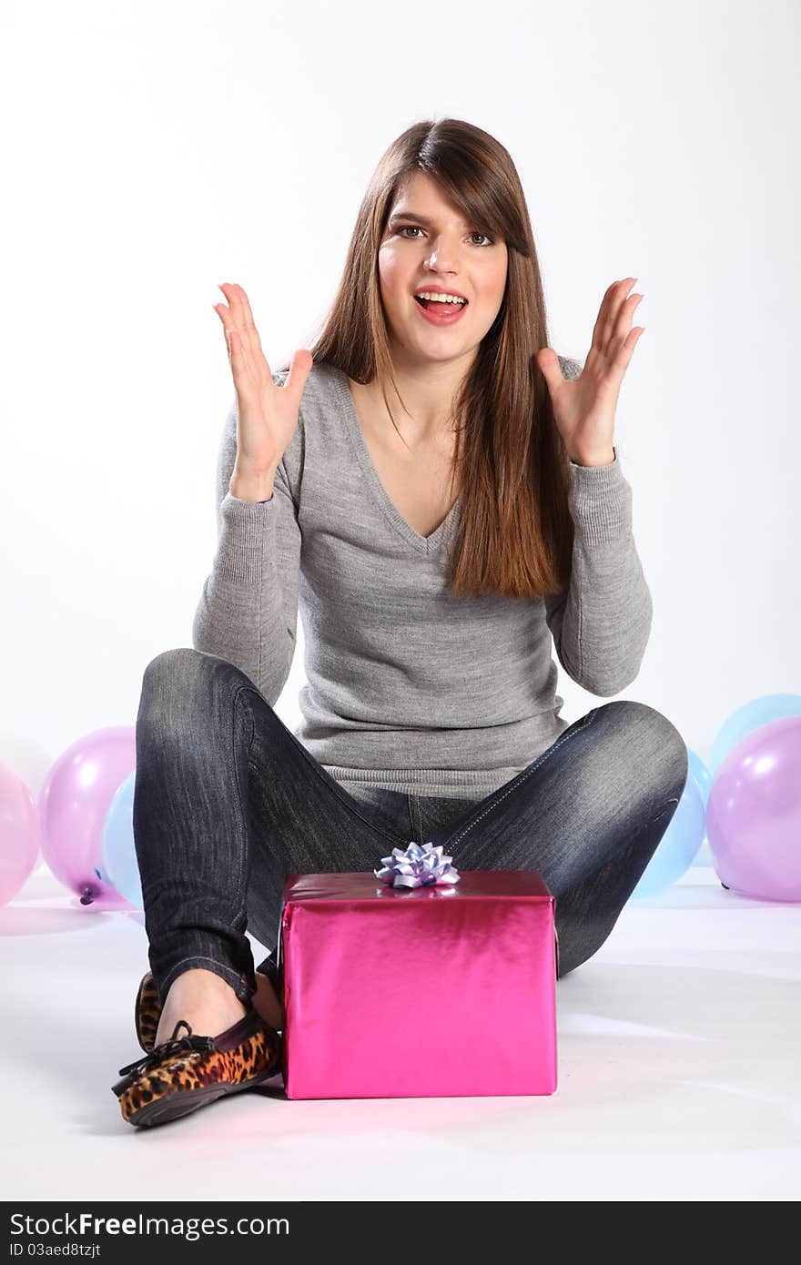 Beautiful young caucasian girl with long hair, has hands raised in happy surprise as she receives a birthday present. Model is wearing jeans and a grey top, sitting on the floor with balloons nearby. Beautiful young caucasian girl with long hair, has hands raised in happy surprise as she receives a birthday present. Model is wearing jeans and a grey top, sitting on the floor with balloons nearby.