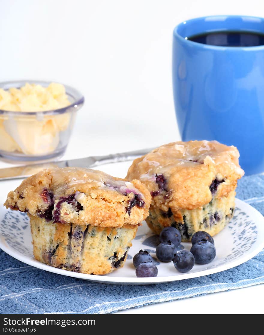 Two blueberry muffins with blueberries on a plate and cup of coffee