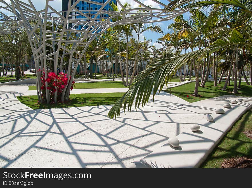 Metal flower sculpture in city park casts a spiderweb shadow on white path and white stone bench with seats separated by polished pebbles. Suitable for concepts dealing with creative use of public spaces. Metal flower sculpture in city park casts a spiderweb shadow on white path and white stone bench with seats separated by polished pebbles. Suitable for concepts dealing with creative use of public spaces.