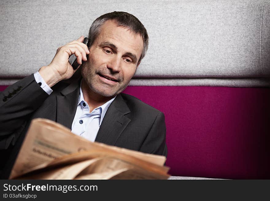 Friendly seniour business executive in grey suit talking on cell phone and reading newspaper. Friendly seniour business executive in grey suit talking on cell phone and reading newspaper.