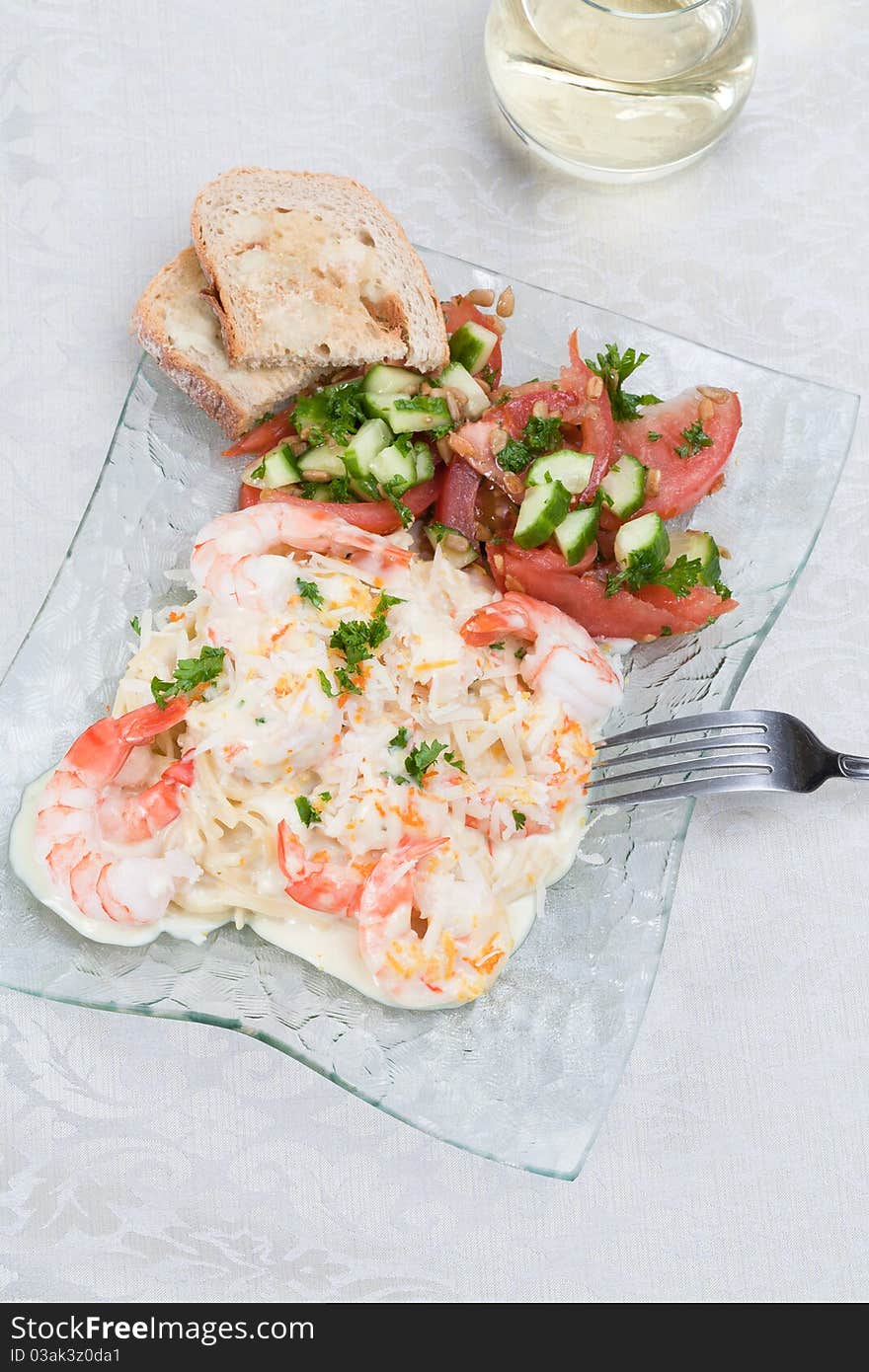 An angled top view of a glass plate of garlic shrimp linguine with a mandarin and garlic sauce with a side of a tomatoes,cucumbers and parlsey salad and garlic butter toast, and a glass of white wine on an off-white linen tablecloth