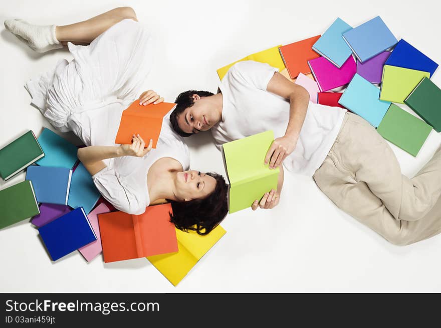 Smiling young man and woman lying on floor with colorful books and reading, looking at books, isolated on white background. Smiling young man and woman lying on floor with colorful books and reading, looking at books, isolated on white background.