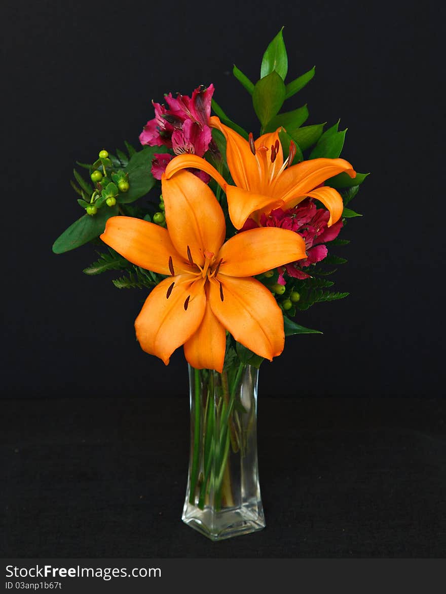 A vibrant bouquet of orange lilies, magenta freesias and greenery in a glass vase, on a black background. A vibrant bouquet of orange lilies, magenta freesias and greenery in a glass vase, on a black background