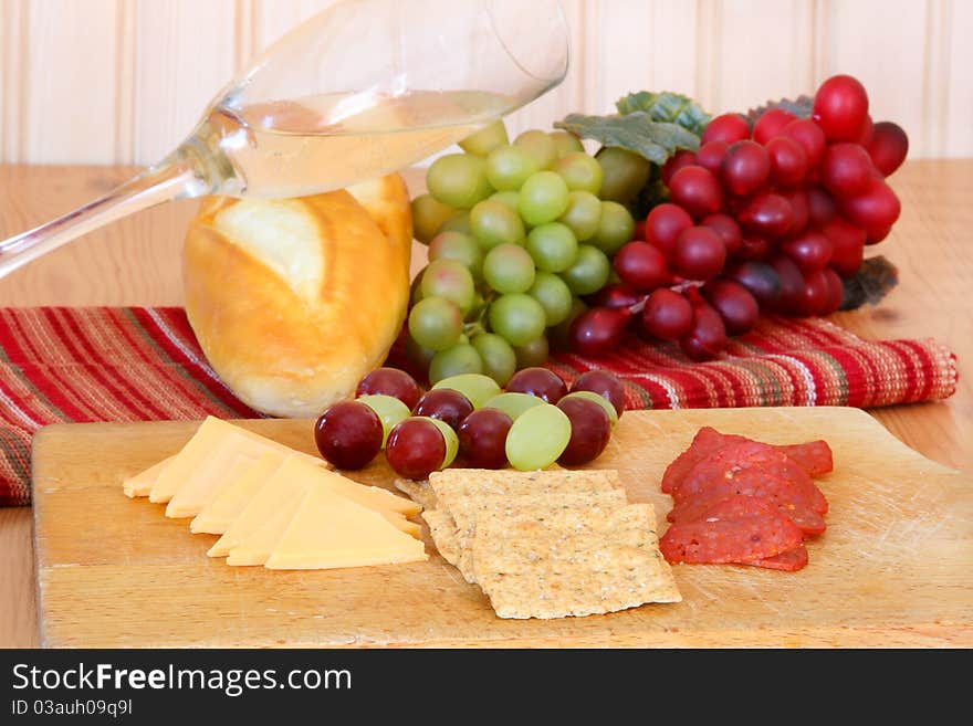 Cheese, crackers, pepperoni, grapes arranged on a cutting board with bread, wine and grapes in the background. Cheese, crackers, pepperoni, grapes arranged on a cutting board with bread, wine and grapes in the background