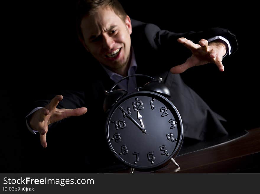 Young desperate businessman in dark suit at office desk jumping at alarm clock showing five minutes to twelve o'clock symbolizing the end is approaching, low-key image isolated on black background. Young desperate businessman in dark suit at office desk jumping at alarm clock showing five minutes to twelve o'clock symbolizing the end is approaching, low-key image isolated on black background.