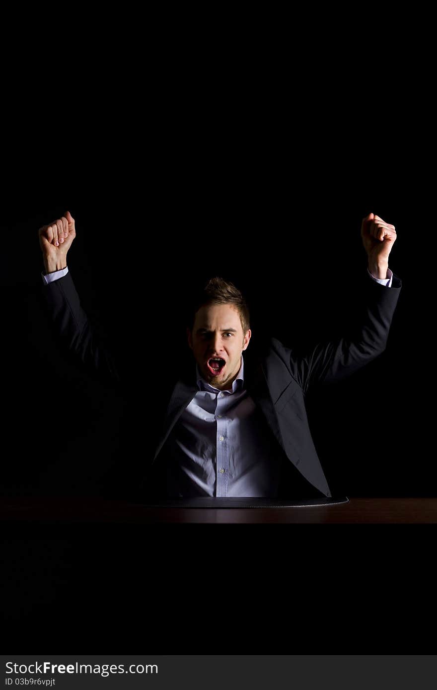 Young modern businessman in dark suit sitting at office desk and raising hands celebrating business success, low-key image isolated on black background. Young modern businessman in dark suit sitting at office desk and raising hands celebrating business success, low-key image isolated on black background.