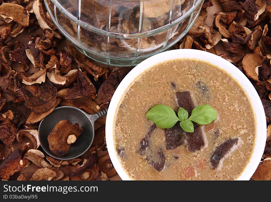 Mushroom soup in a bowl, decorated with basil, with dried mushrooms surrounding it and some in a jar, and an antique spoon