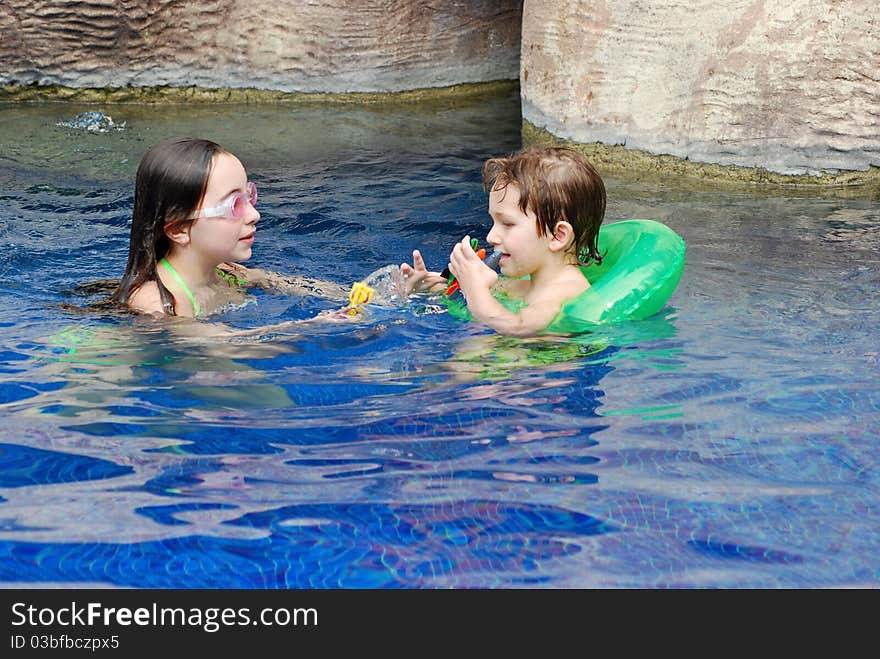 A boy and girl, brother and sister, in the pool playing together while the younger one wears a green float. A boy and girl, brother and sister, in the pool playing together while the younger one wears a green float.