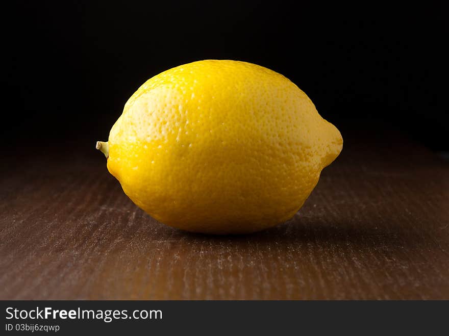 A single fresh yellow lemon on a wood grain table against a black background. A single fresh yellow lemon on a wood grain table against a black background.