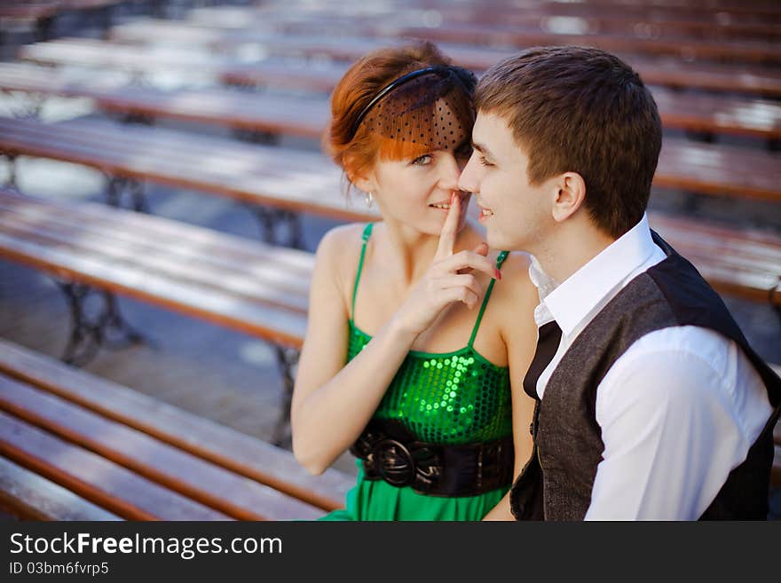 Closeup portraits of young couple sitting together on park bench. Girl making a keep it quiet. Focus on a guy