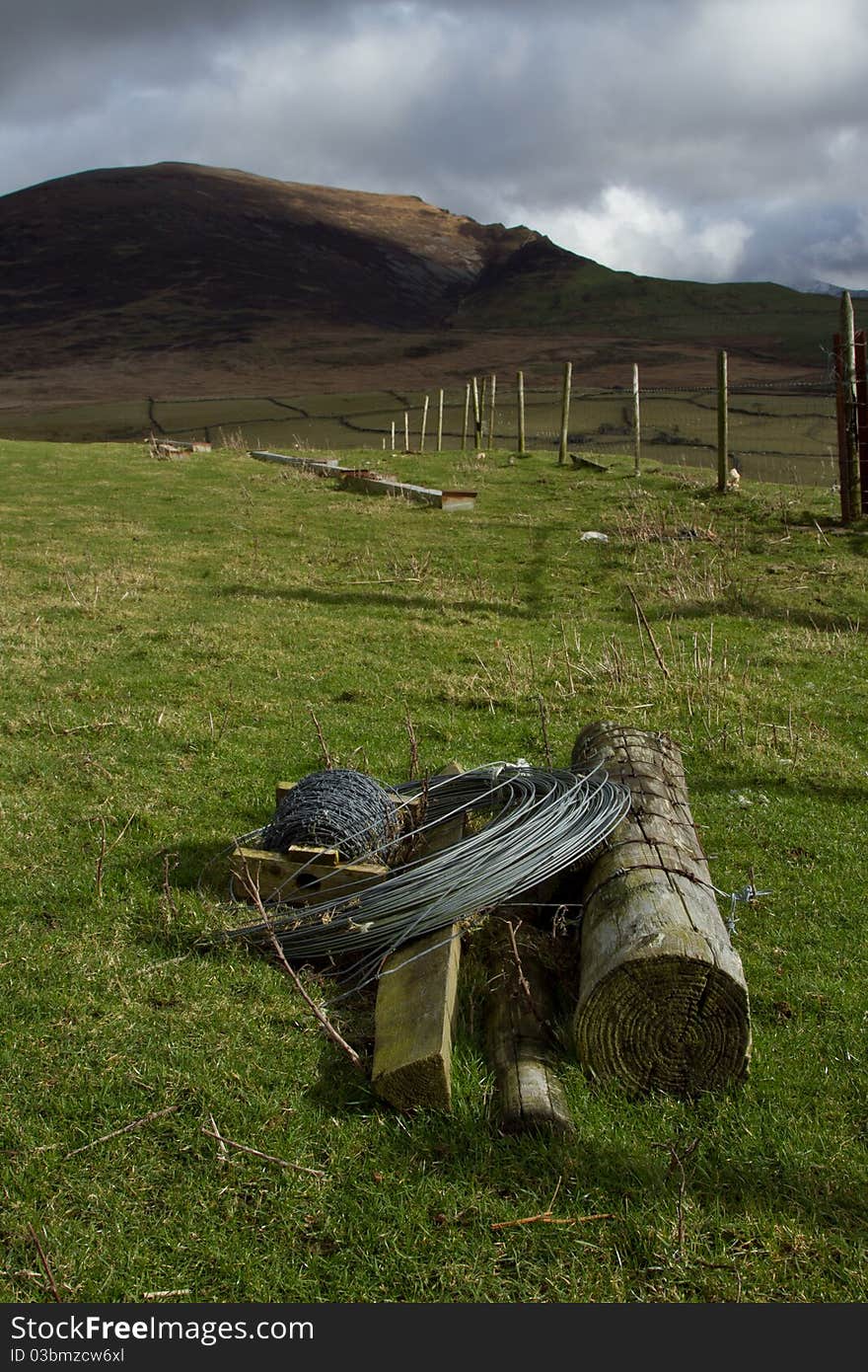 Agricultural landscape with wooden fence posts a coil of wire and a spool of barbed wire on green grass with a hill in the background. Agricultural landscape with wooden fence posts a coil of wire and a spool of barbed wire on green grass with a hill in the background.