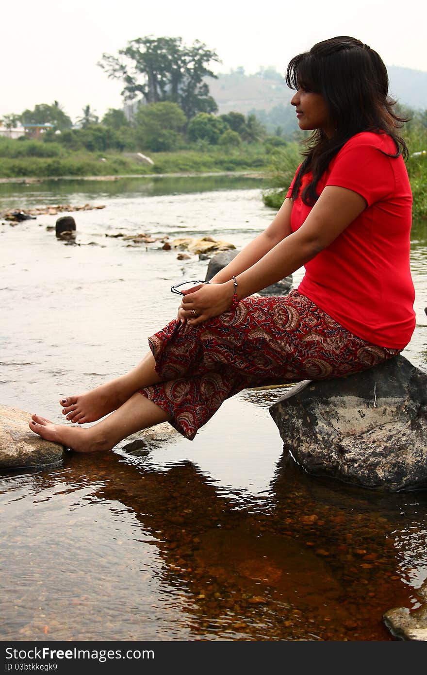 Young indian woman in casual red dress sitting on a rock in water. Young indian woman in casual red dress sitting on a rock in water