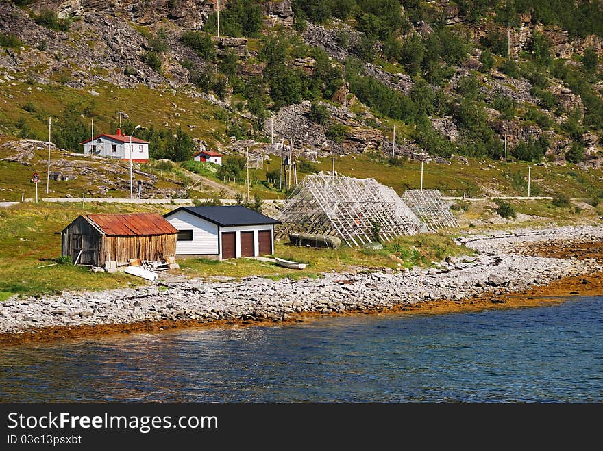 Norwegian shore is photographed in summer. There are some wooden houses and sheds under moss mountain, scaffoldings for dried cod near edge of quite water. Norwegian shore is photographed in summer. There are some wooden houses and sheds under moss mountain, scaffoldings for dried cod near edge of quite water.