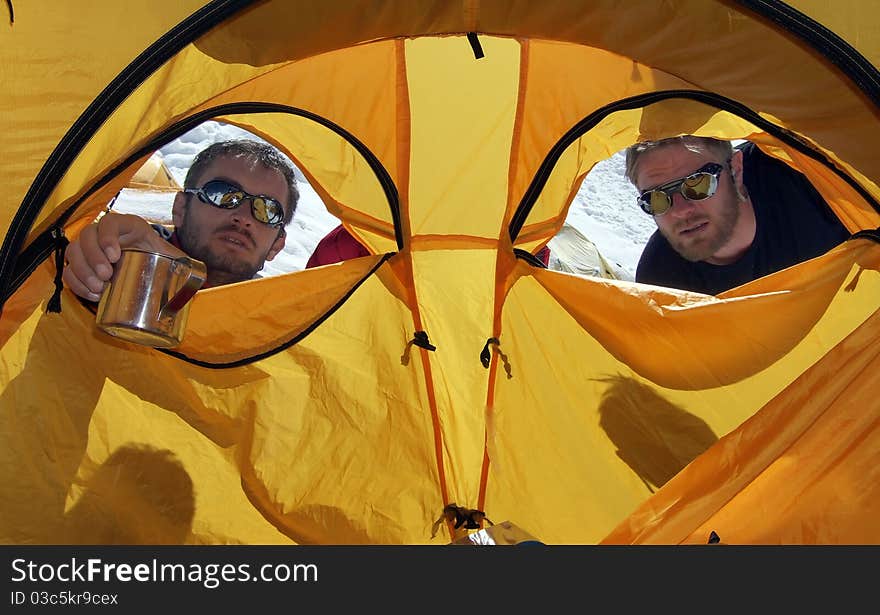 Two men are staring at the third one, situated in a tent, in a snowy environment. A metalic cup of tea is offered to him. Two men are staring at the third one, situated in a tent, in a snowy environment. A metalic cup of tea is offered to him.